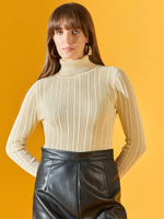 Woman wears 98degreenorth cream ribbed turtleneck sweater and black leather skirt against a yellow background.