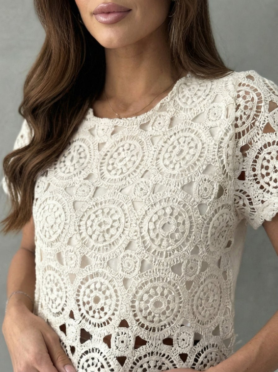 Woman in 98degreenorth Women's Crochet Lace Crop Top—off white, short sleeves, scalloped hem—against a gray background.
