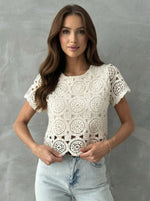 Woman in 98degreenorth Women's Crochet Lace Crop Top—Off White, short sleeve, scalloped hem, stands by gray wall.
