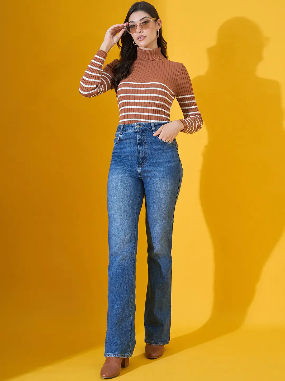 Woman wears 98degreenorth Women’s Dark Camel Ribbed Turtleneck Sweater, posing against a yellow background.