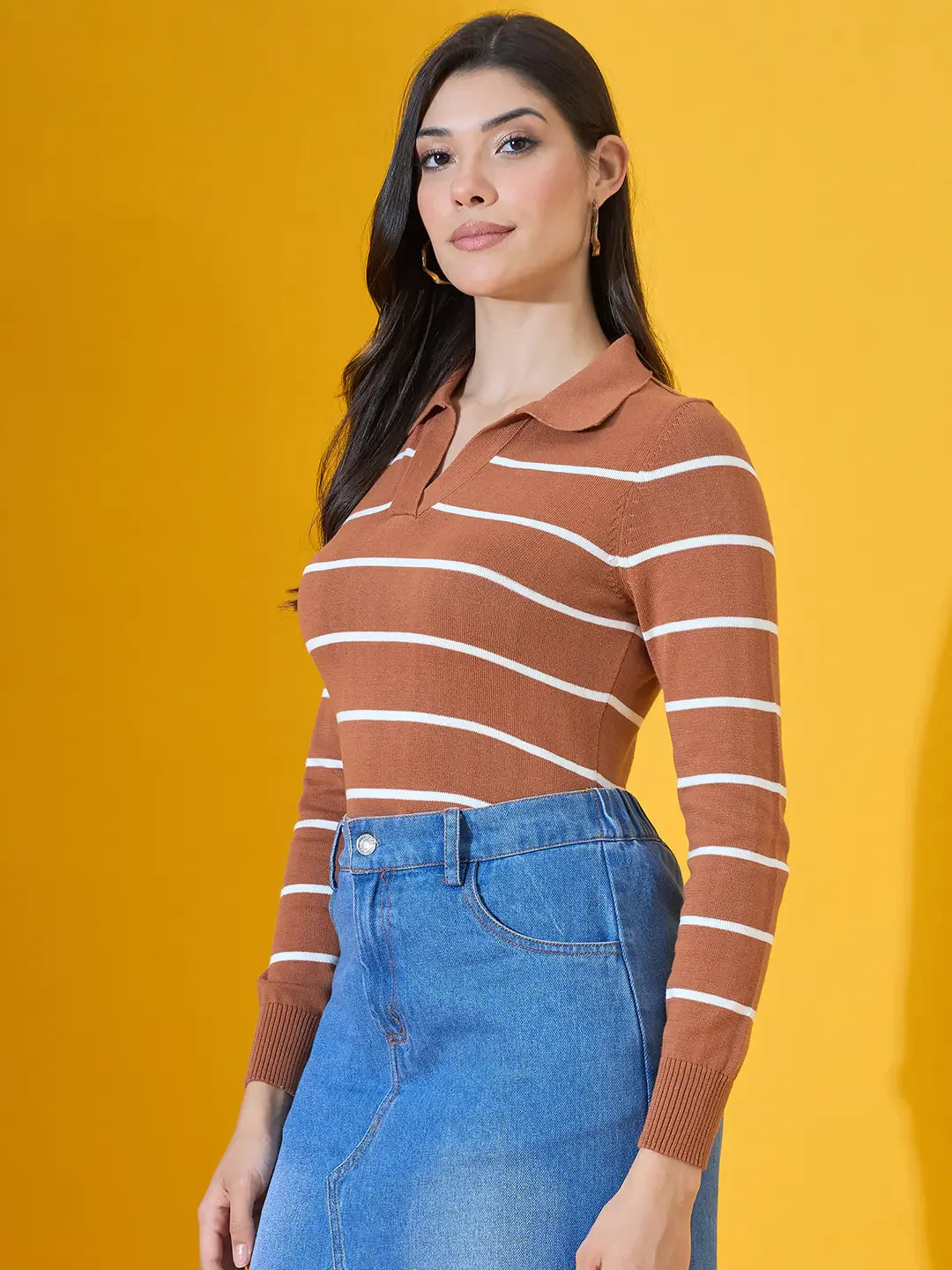 Woman in a 98degreenorth dark camel striped collared sweater and blue denim skirt stands before a yellow background.