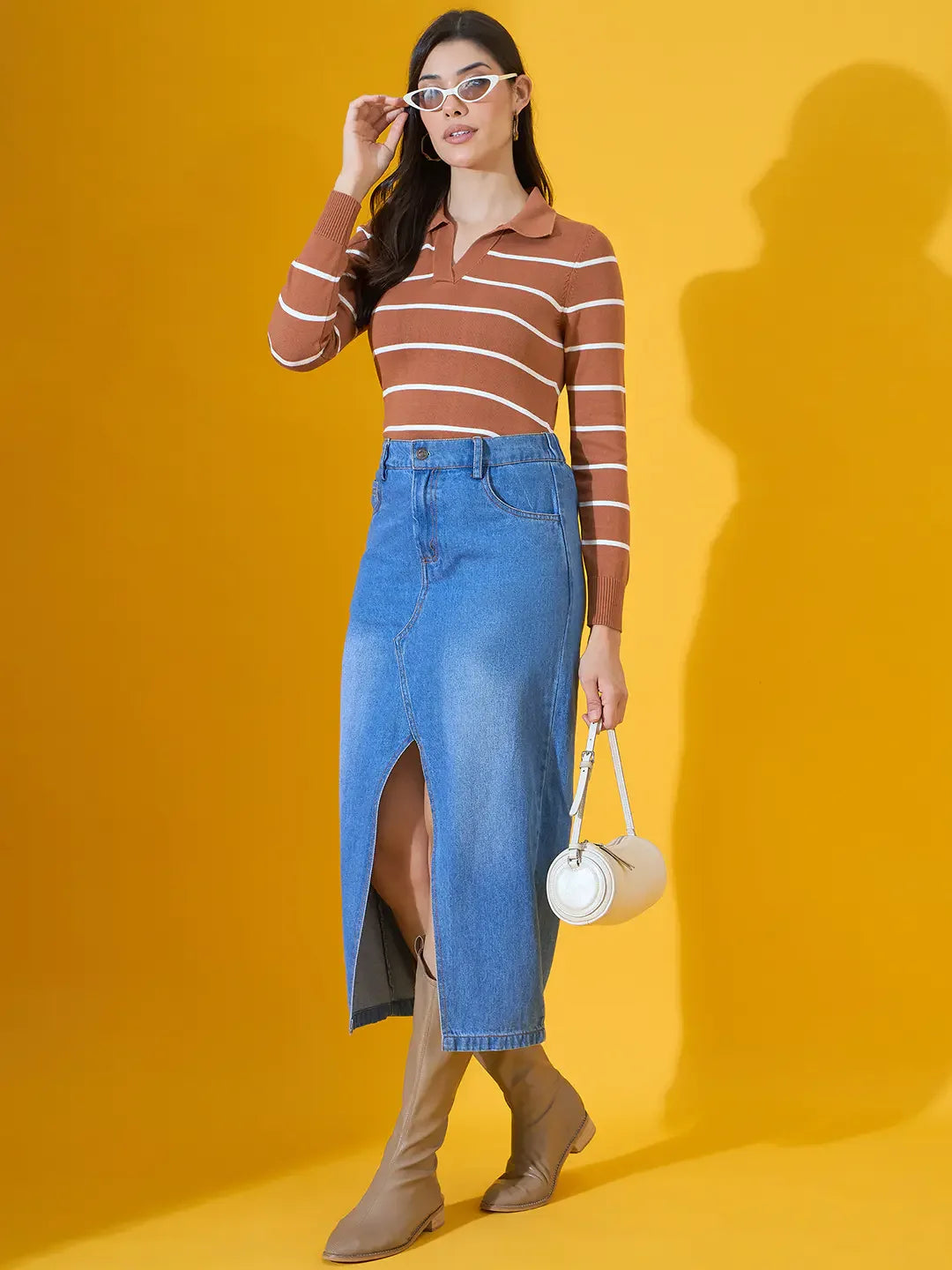 Woman wears 98degreenorth dark camel striped collared sweater, denim skirt, tan boots, and white sunglasses on yellow.