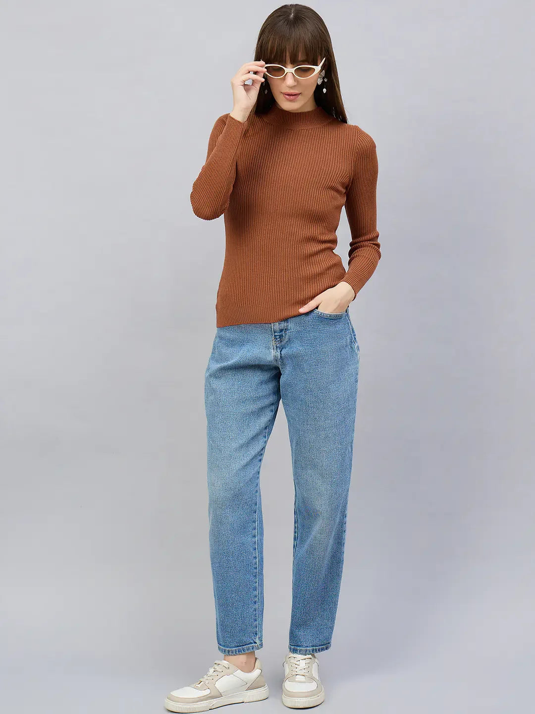 Woman in 98degreenorth dark camel turtle neck sweater with jeans and sneakers, adjusting sunglasses against grey backdrop.