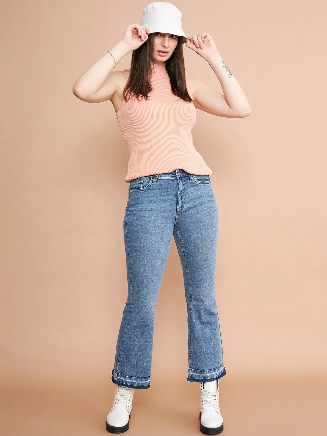 Woman in a 98degreenorth dark pink ribbed high neck sleeveless knit top, blue jeans, and white boots poses by beige wall.