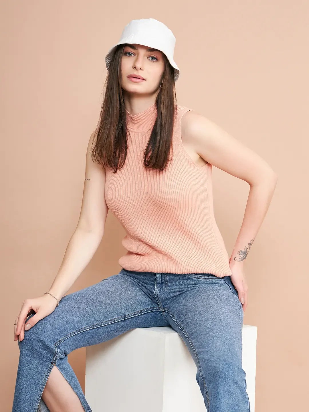 Woman in a 98degreenorth dark pink ribbed high neck sleeveless top sits on a white block against a beige background.