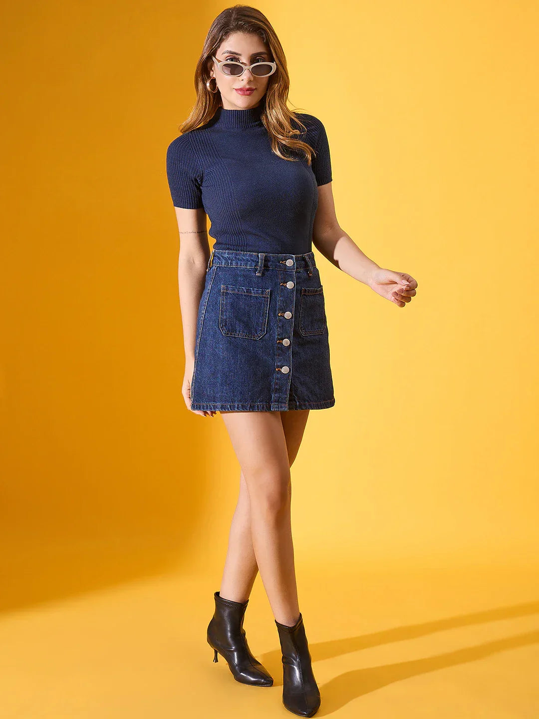 Woman models 98degreenorth blue ribbed turtleneck sweater and denim skirt against a yellow background.