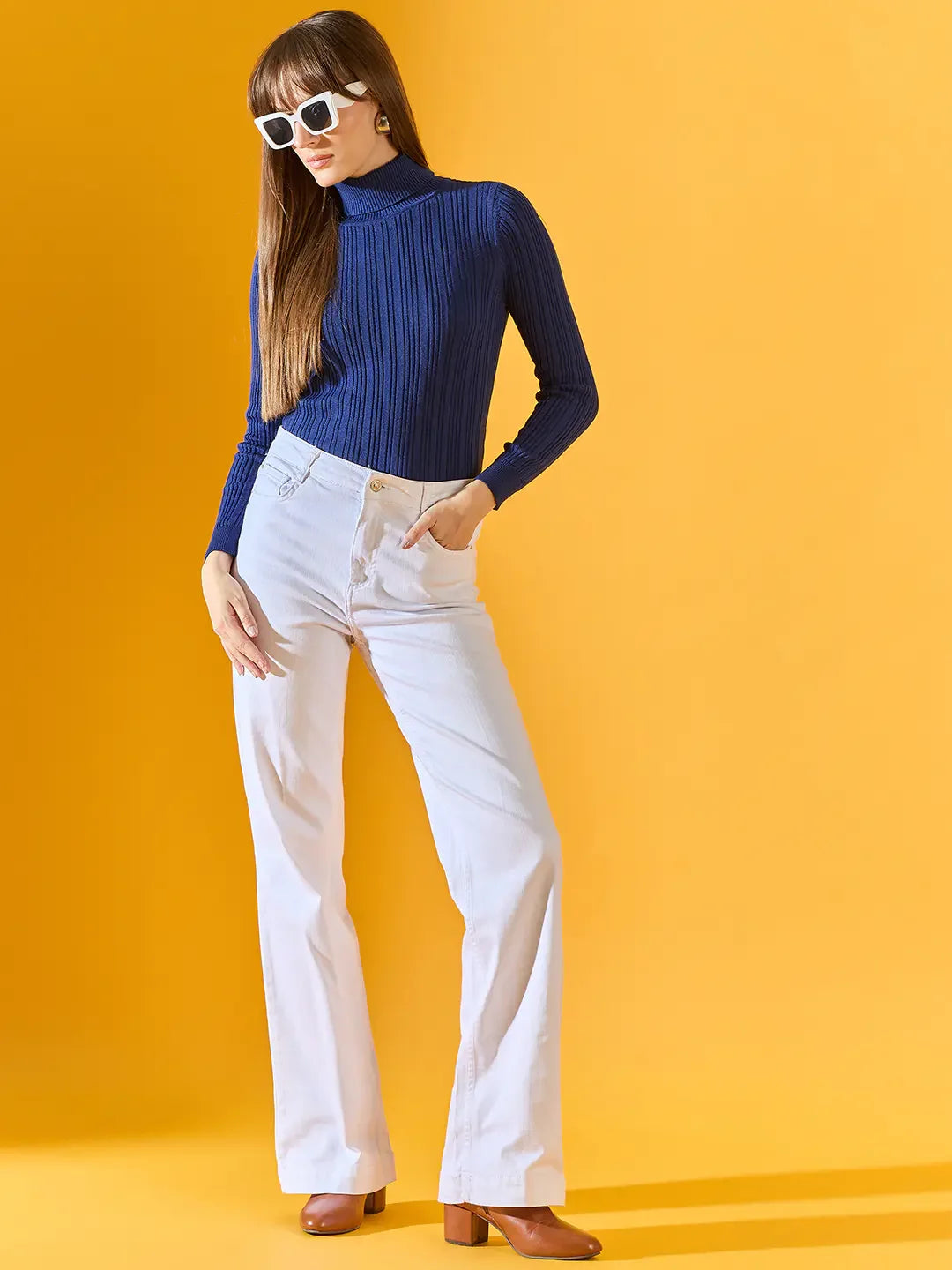 Woman in 98degreenorth denim blue ribbed turtleneck sweater, white pants, and heels against a bright yellow background.