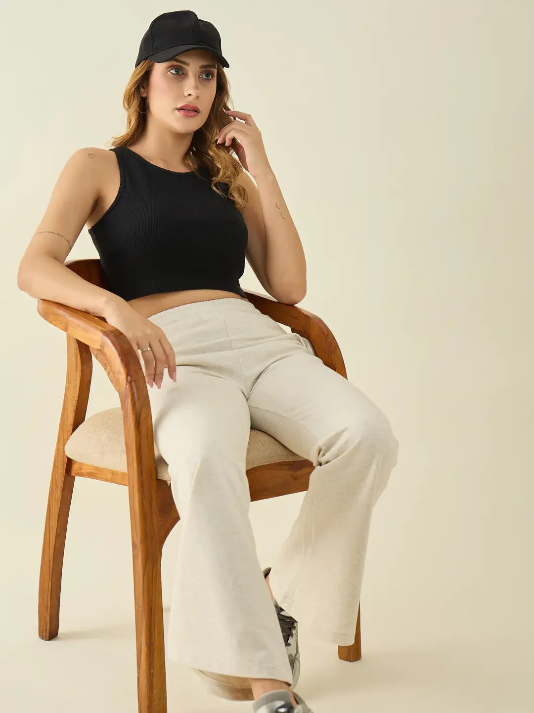 Woman in a black crop top, cap, and 98degreenorth ecru flared bootcut stretchable trousers sits on a wooden chair.