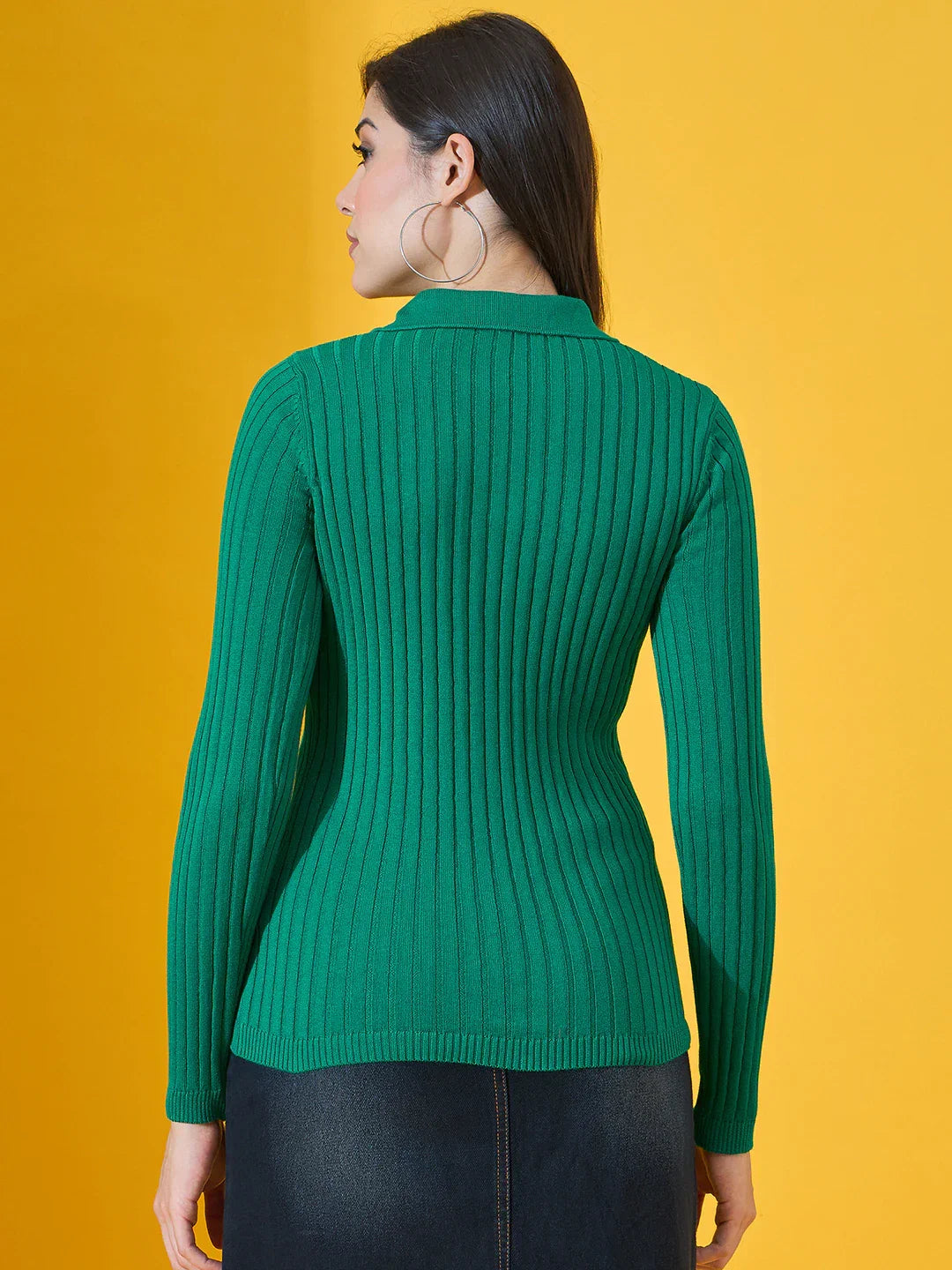 A woman in a 98degreenorth emerald green ribbed collar neck sweater and hoop earrings stands against a yellow background.