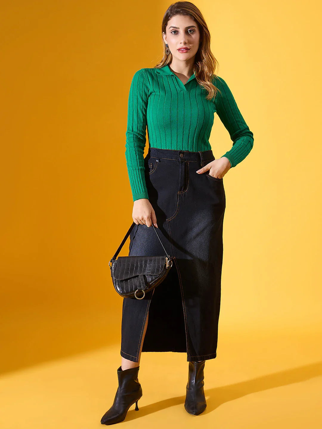 Woman in a 98degreenorth emerald green ribbed collar sweater and black skirt holds a black bag against a yellow backdrop.