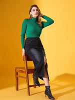 Woman in 98degreenorth's emerald green ribbed turtle neck sweater and black skirt sits on a chair, wearing black boots.