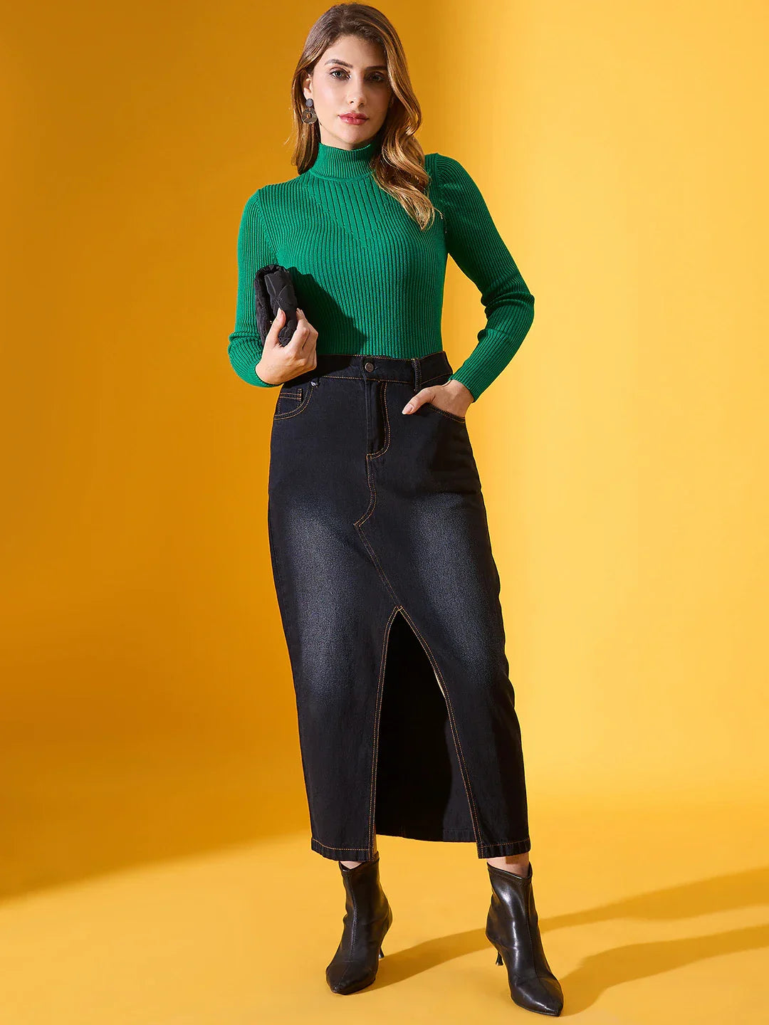 Woman wears 98degreenorth’s emerald green ribbed turtleneck sweater and black skirt, holding a clutch against yellow.