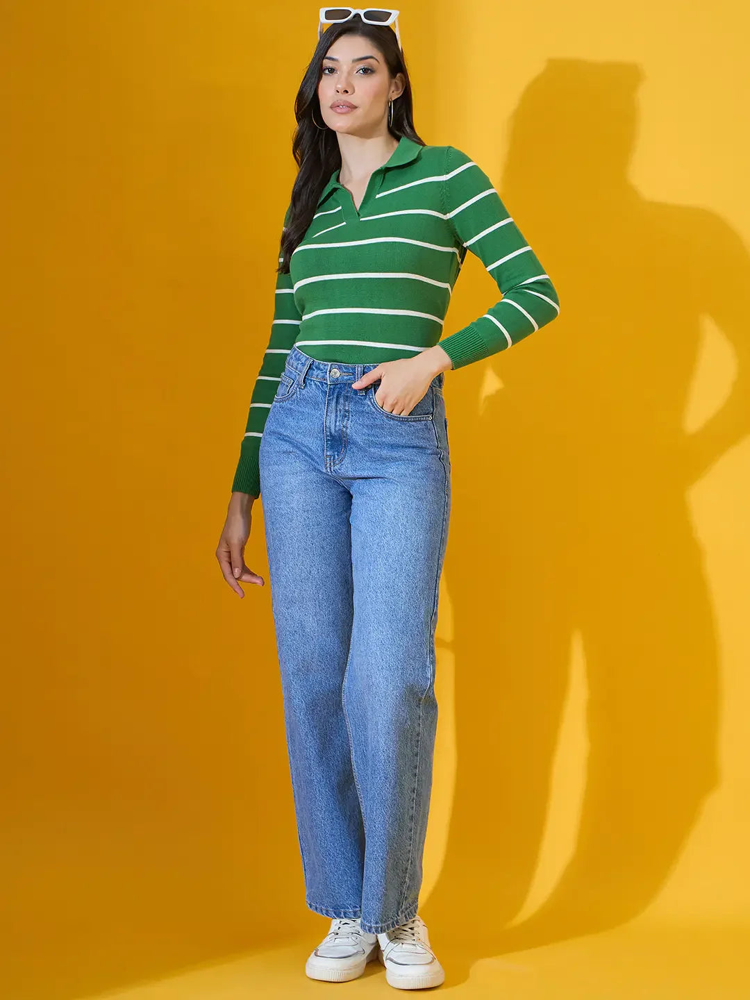 A woman wears a 98degreenorth fern green striped collared sweater with blue jeans and white sneakers against a yellow background.