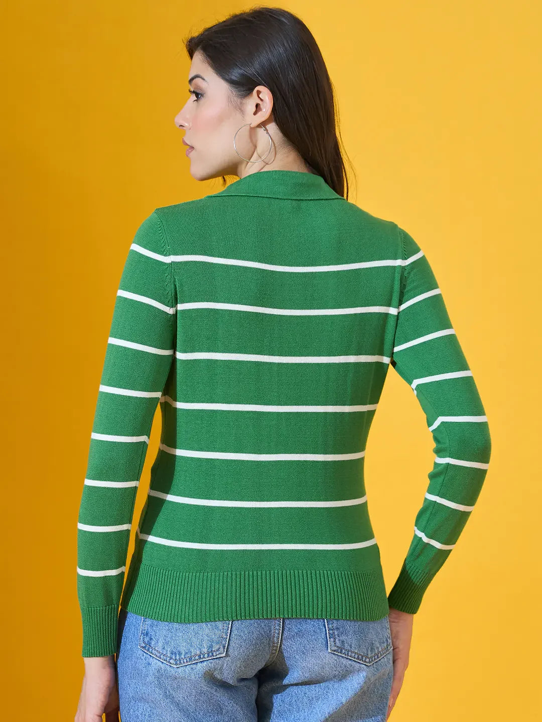 Woman in a 98degreenorth fern green striped collared sweater, blue jeans, facing away against a yellow background.