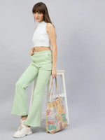 Woman in a white crop top and 98degreenorth’s Pista flared fleece sweatpants stands by a white stool with a tote bag.