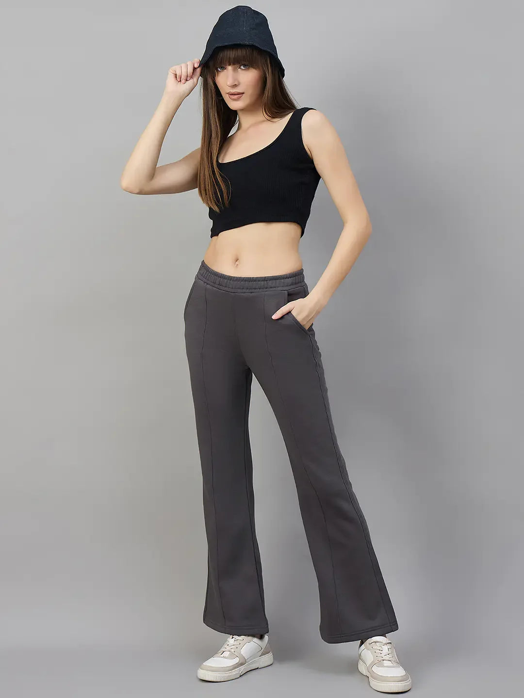 Woman in a black crop top, 98degreenorth Flared Sweatpants in Charcoal Grey, white sneakers, and bucket hat poses.