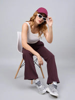 Woman in white tank, 98degreenorth Dark Wine Flared Sweatpants, maroon cap & sunglasses sits on a chair.