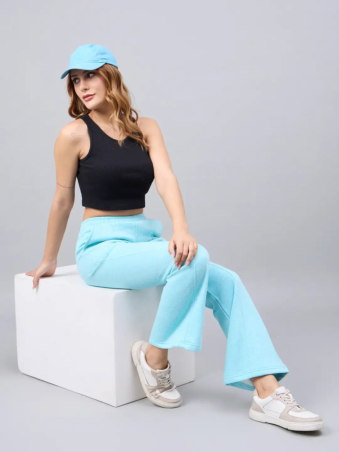 Woman wears 98degreenorth Women’s Flared Sweatpants in Turquoise Blue, crop top, and cap on white cube against gray backdrop.