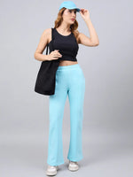 Woman in black crop top, 98degreenorth turquoise flared sweatpants, blue cap & white sneakers on gray background.