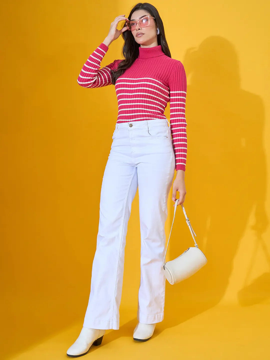 Woman in a 98degreenorth fuchsia ribbed knit turtleneck sweater poses with a white purse against a yellow background.