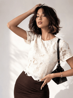 Woman in a beige 98degreenorth lace overlay crop top and brown skirt poses, holding a black handbag and touching her hair.