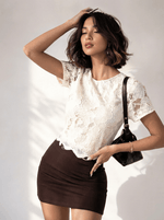 Woman wears 98degreenorth's beige floral lace overlay crop top and dark brown skirt, holding a black bag.