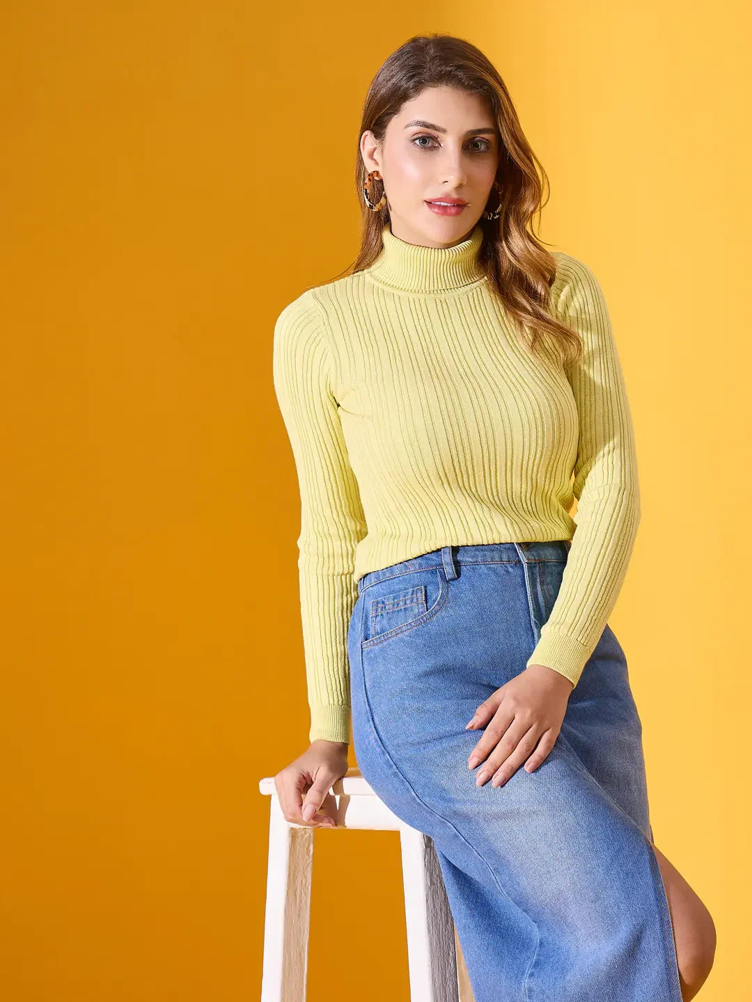 Woman in 98degreenorth Lemon Ribbed Turtleneck Sweater and blue skirt sits on a white stool, yellow background.