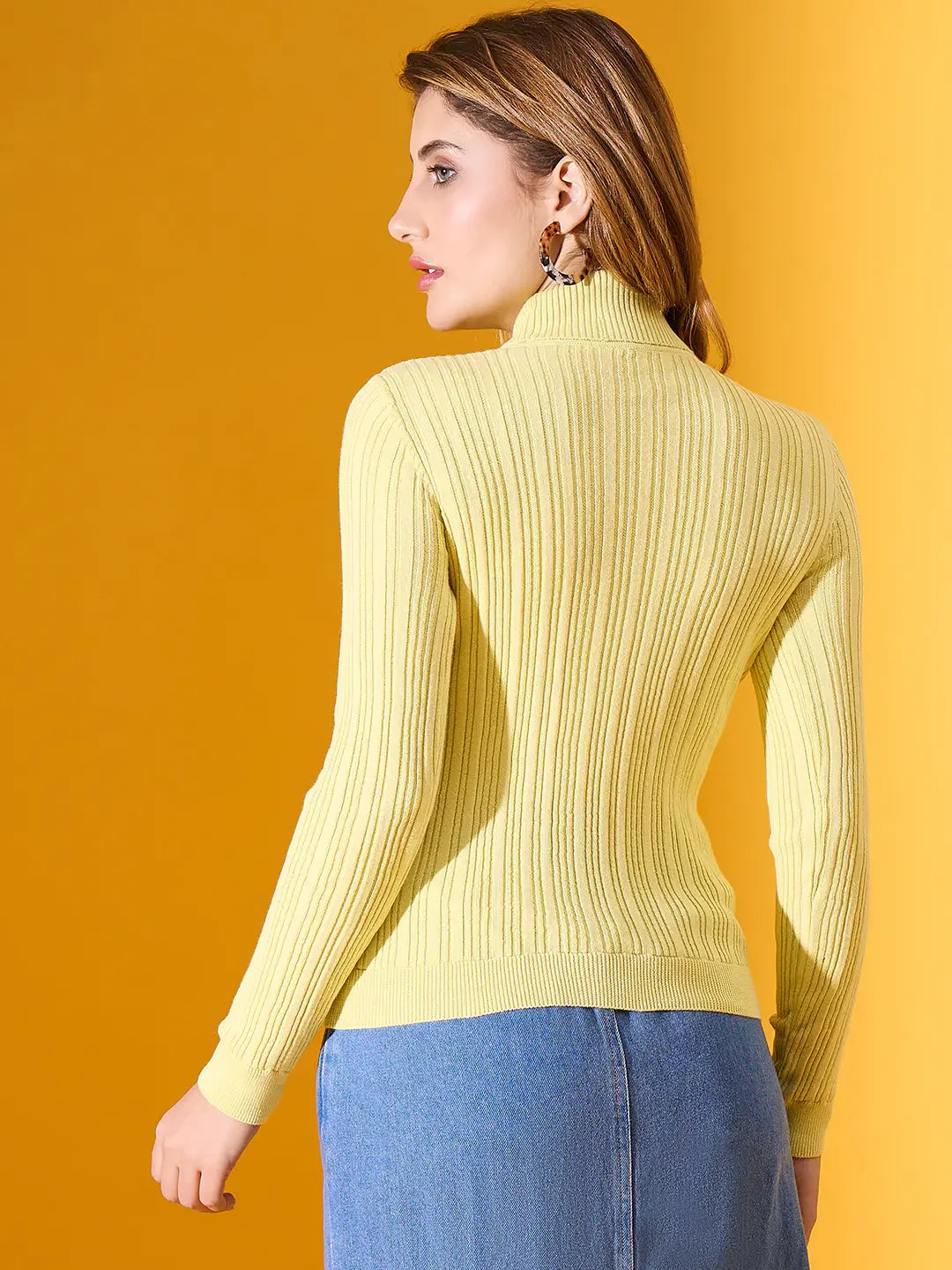 Woman in a 98degreenorth Lemon Ribbed Turtleneck Sweater and blue skirt stands against a yellow background.