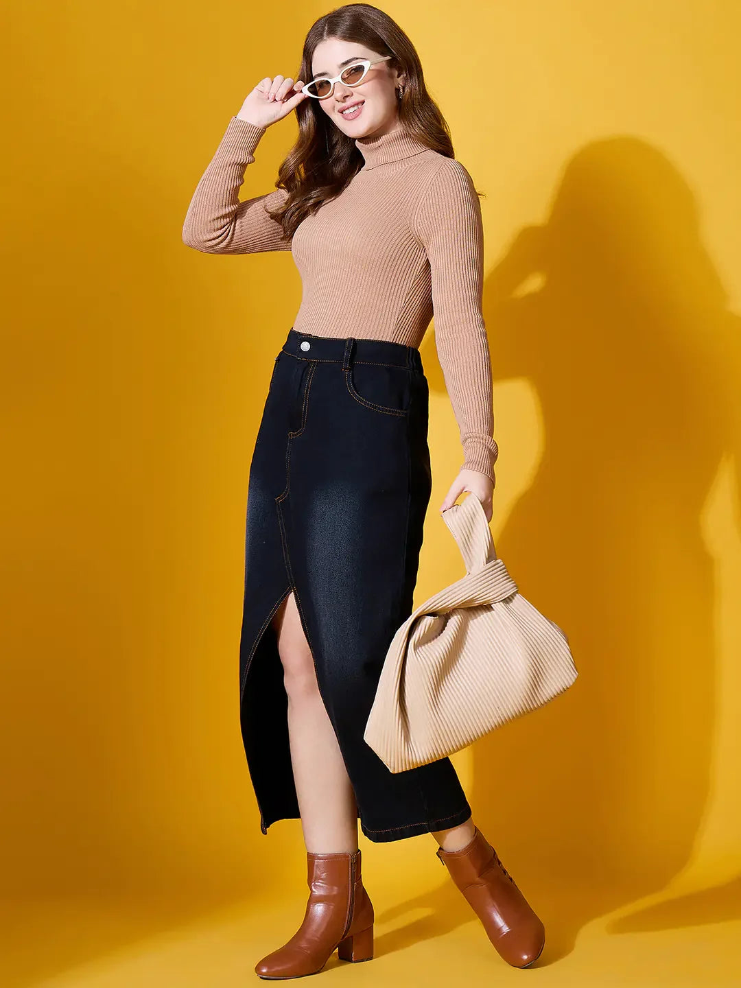 Woman in a 98degreenorth Light Camel High Turtle-Neck Ribbed Sweater, black skirt, brown boots, with pleated bag.