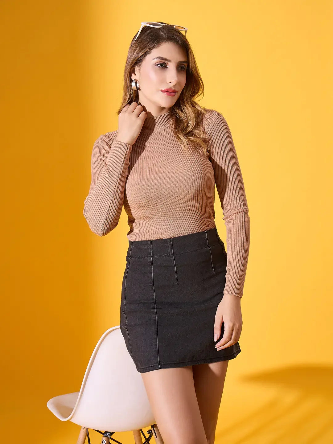 Woman in 98degreenorth’s light camel ribbed high-neck pullover and black skirt poses by a white chair on yellow background.