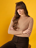Woman in a 98degreenorth light camel ribbed turtle neck slim fit sweater sits with arms crossed against a yellow backdrop.
