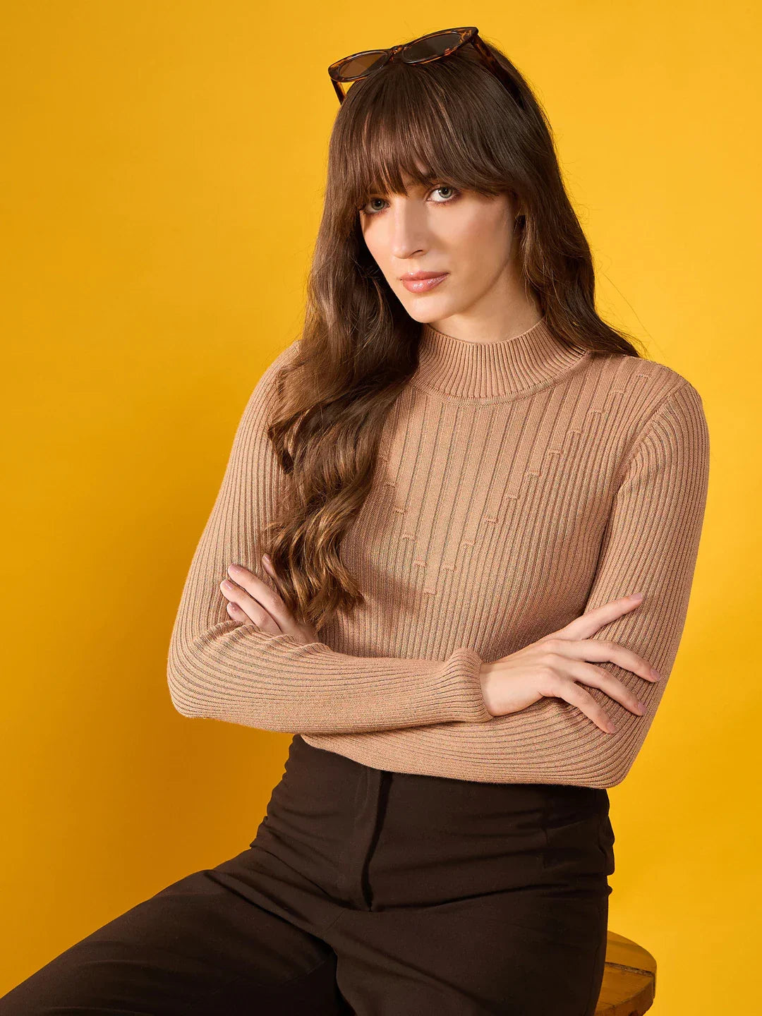 Woman in a 98degreenorth light camel ribbed turtle neck slim fit sweater sits with arms crossed against a yellow backdrop.