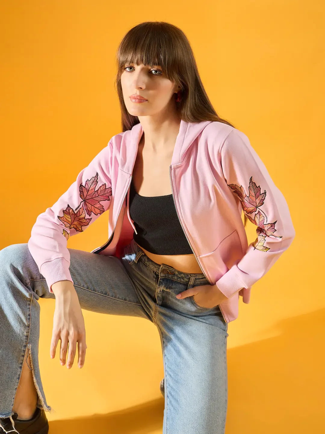 Woman in a 98degreenorth light pink fleece hoodie with leaf embroidery poses against a vibrant yellow background.