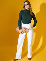 Woman in a 98degreenorth military green ribbed turtleneck sweater, white pants, brown shoes, sunglasses, and beige clutch.