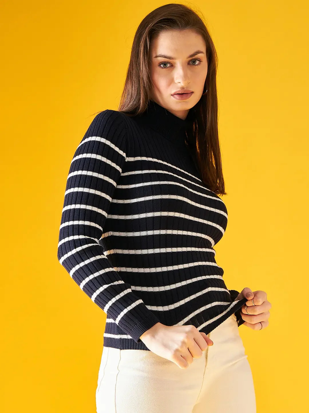 Woman wears 98degreenorth Women’s Navy Blue Striped Ribbed Turtleneck Sweater with white pants on a yellow backdrop.