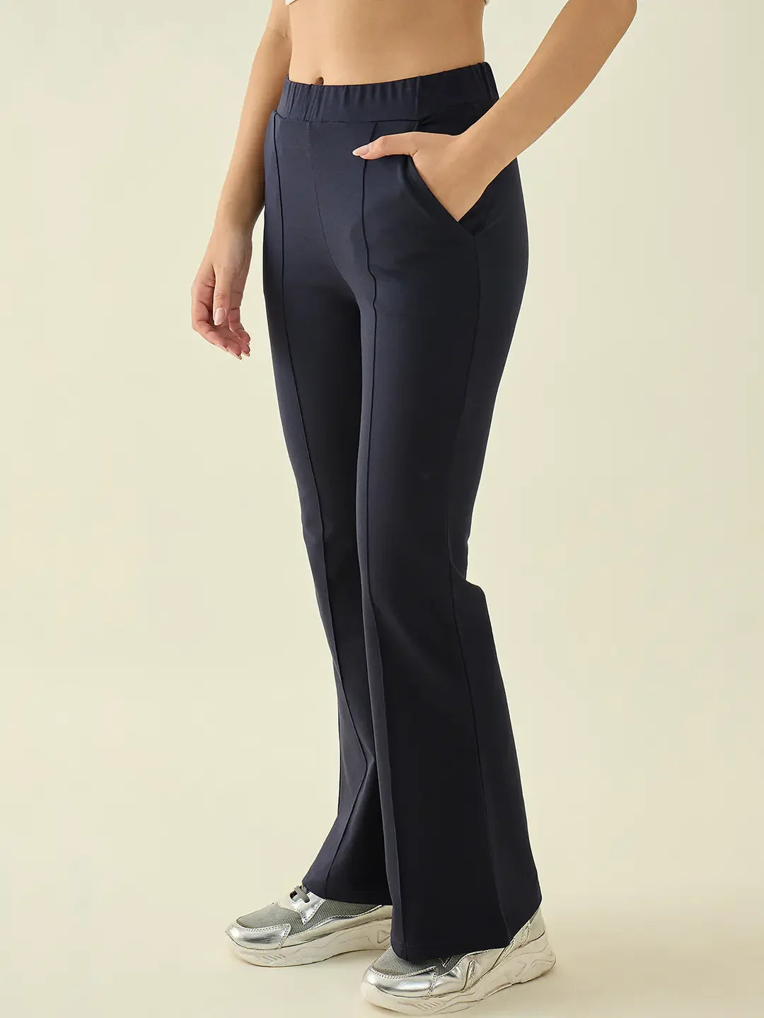 A woman wears 98degreenorth navy flared bootcut cotton trousers and silver sneakers, standing against a beige backdrop.