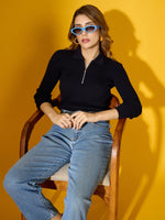 Woman in 98degreenorth’s navy ribbed collar zipper sweater sits on a wooden chair against a yellow background.
