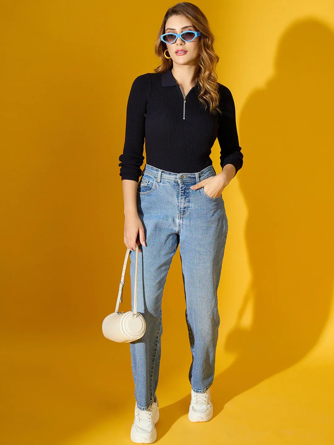 Woman in 98degreenorth Navy Ribbed Collar Zipper Sweater, blue jeans, and white bag poses on a yellow background.