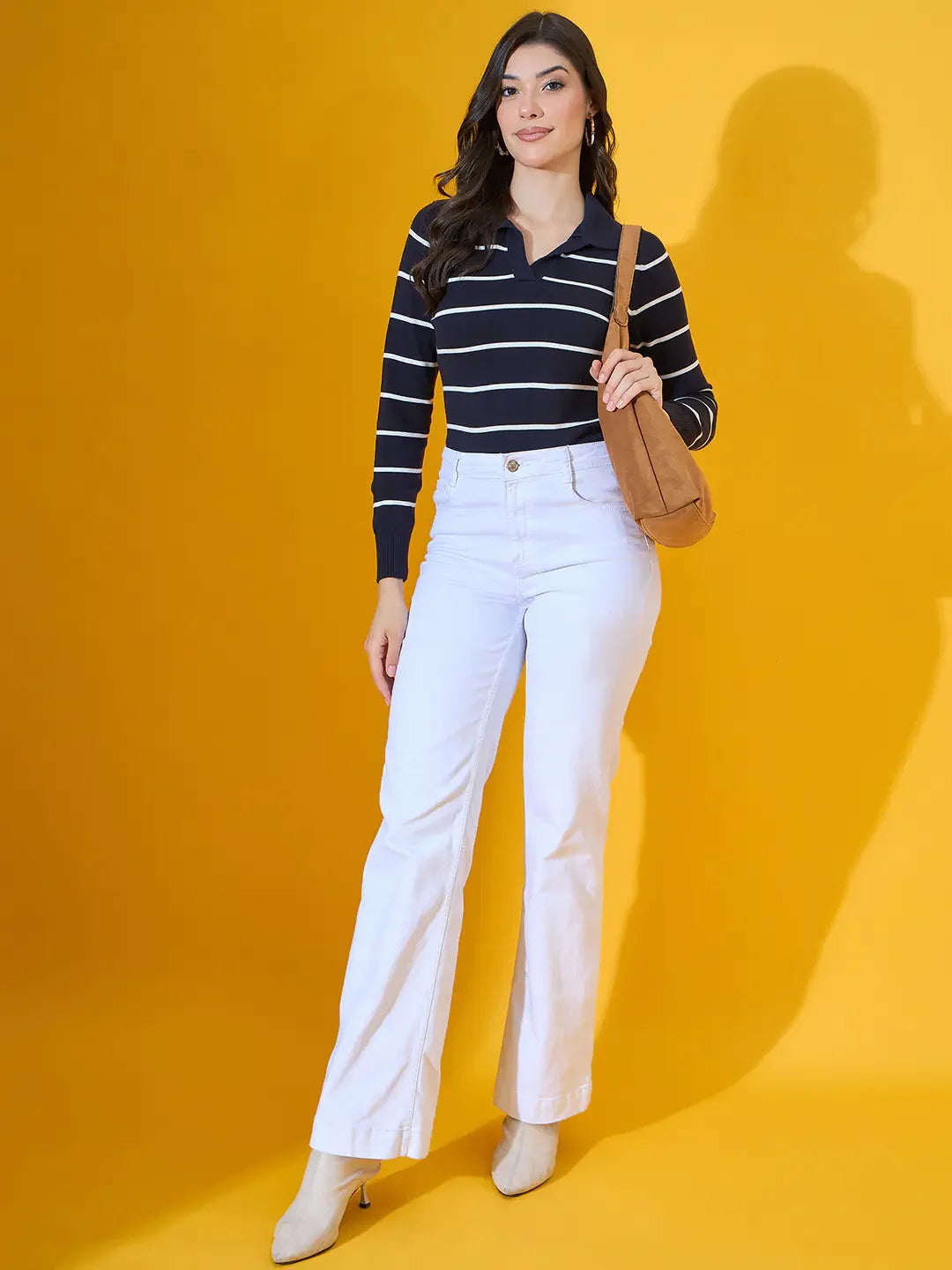 A woman in white pants and boots models a 98degreenorth navy striped collared knit sweater with a brown bag.