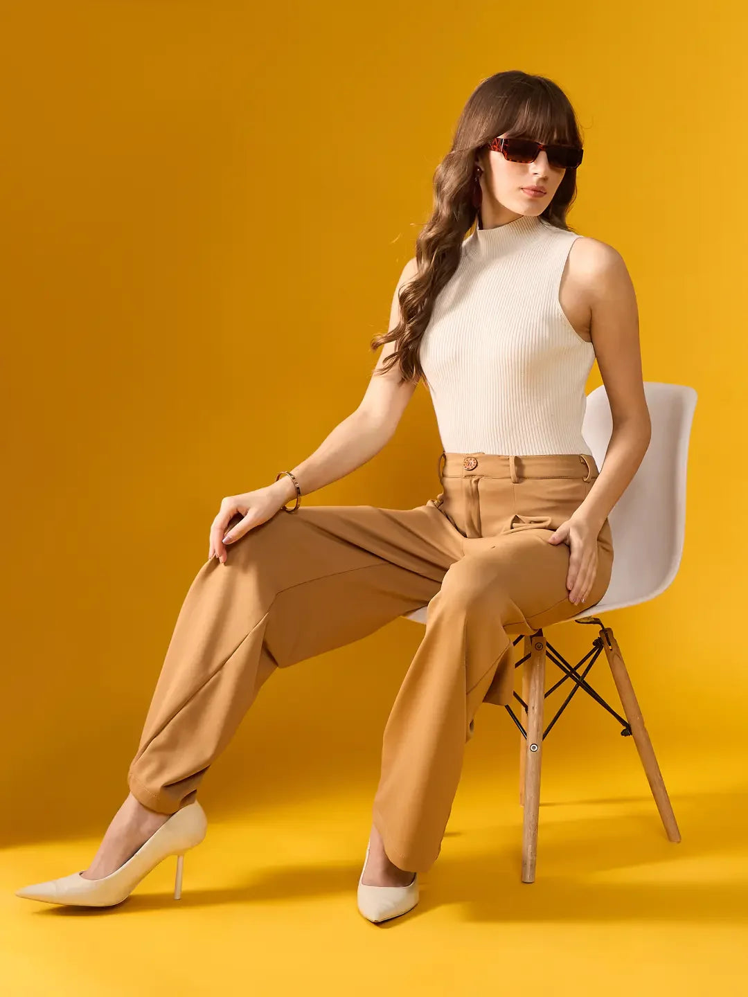 Woman in 98degreenorth Off-White Ribbed High Neck Knit Tank sits on white chair against yellow backdrop, wearing sunglasses.