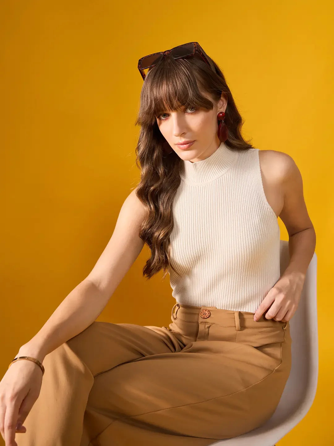 Woman wears 98degreenorth Off-White Ribbed High Neck Sleeveless Knit Top and tan pants, seated by a yellow background.