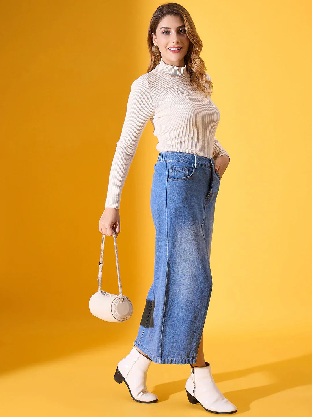 Woman in a 98degreenorth off white ribbed turtleneck sweater, blue denim skirt, and white boots stands by a yellow backdrop.