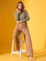 Woman in 98degreenorth pista melange ribbed collar sweater and tan pants sits on stool against yellow backdrop, in white heels.