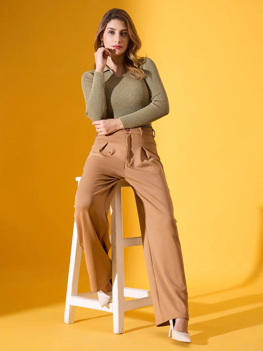 Woman in 98degreenorth pista melange ribbed collar sweater and tan pants sits on stool against yellow backdrop, in white heels.