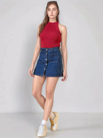 Woman in a 98degreenorth red ribbed halter neck knit top and denim skirt stands against a gray wall.