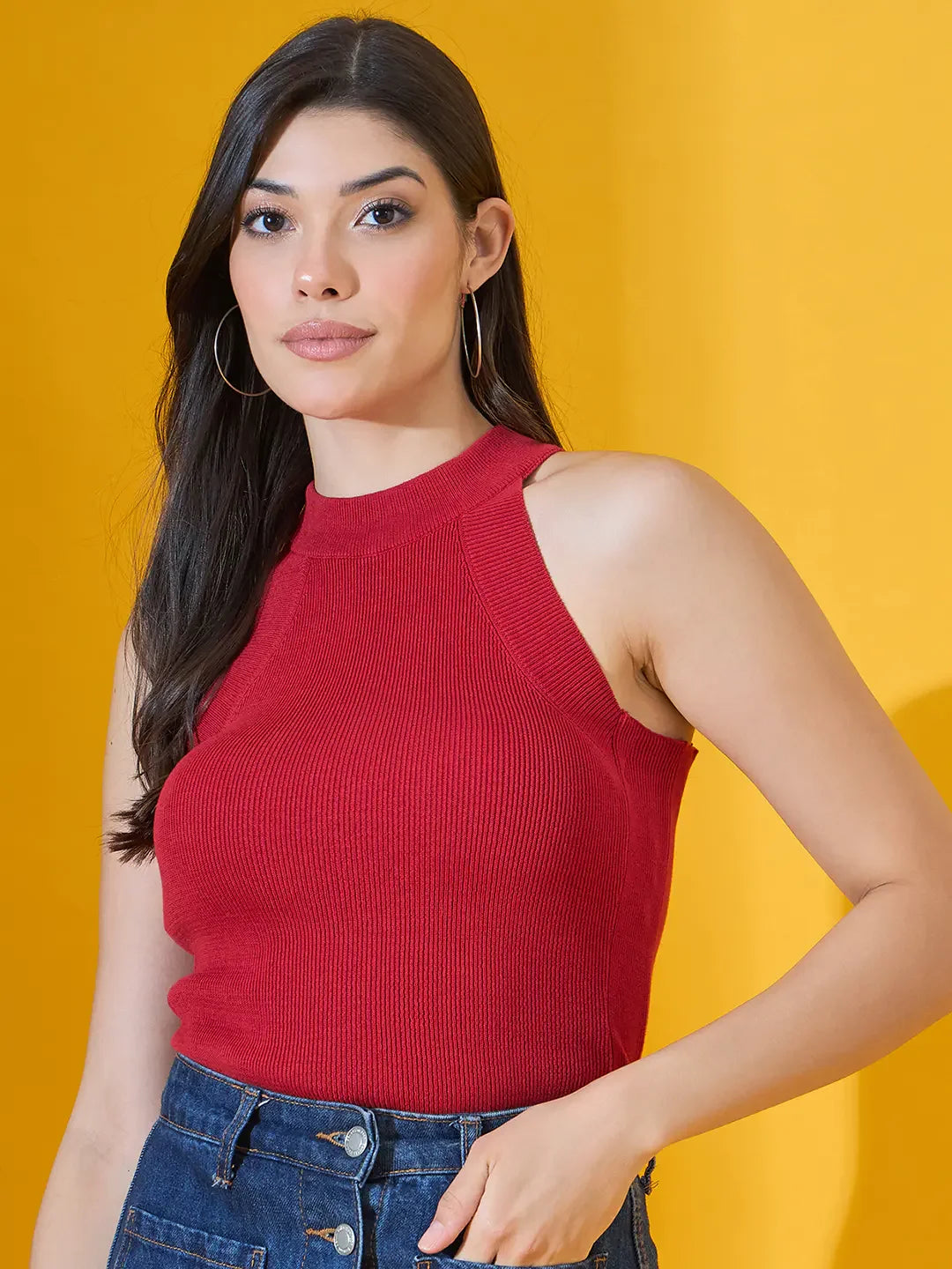 A woman wears the 98degreenorth Red Ribbed Halter Neck Sleeveless Knit Top with blue jeans against a yellow background.