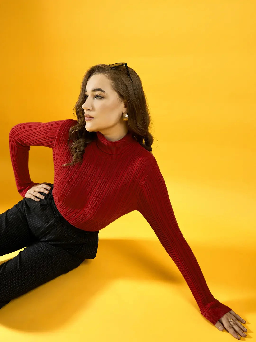 Woman in a 98degreenorth red ribbed turtleneck sweater and black pants sits on a yellow background, looking aside.