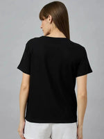 Woman with long brown hair in a 98degreenorth Typography Print Cotton T-shirt and white shorts, seen from the back.