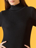 Woman in 98degreenorth Women’s Ribbed Knitted Bodycon Dress – Black, hands on hips, yellow background.