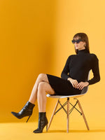Woman wears 98degreenorth black ribbed turtleneck bodycon dress and sunglasses, seated against a yellow background.